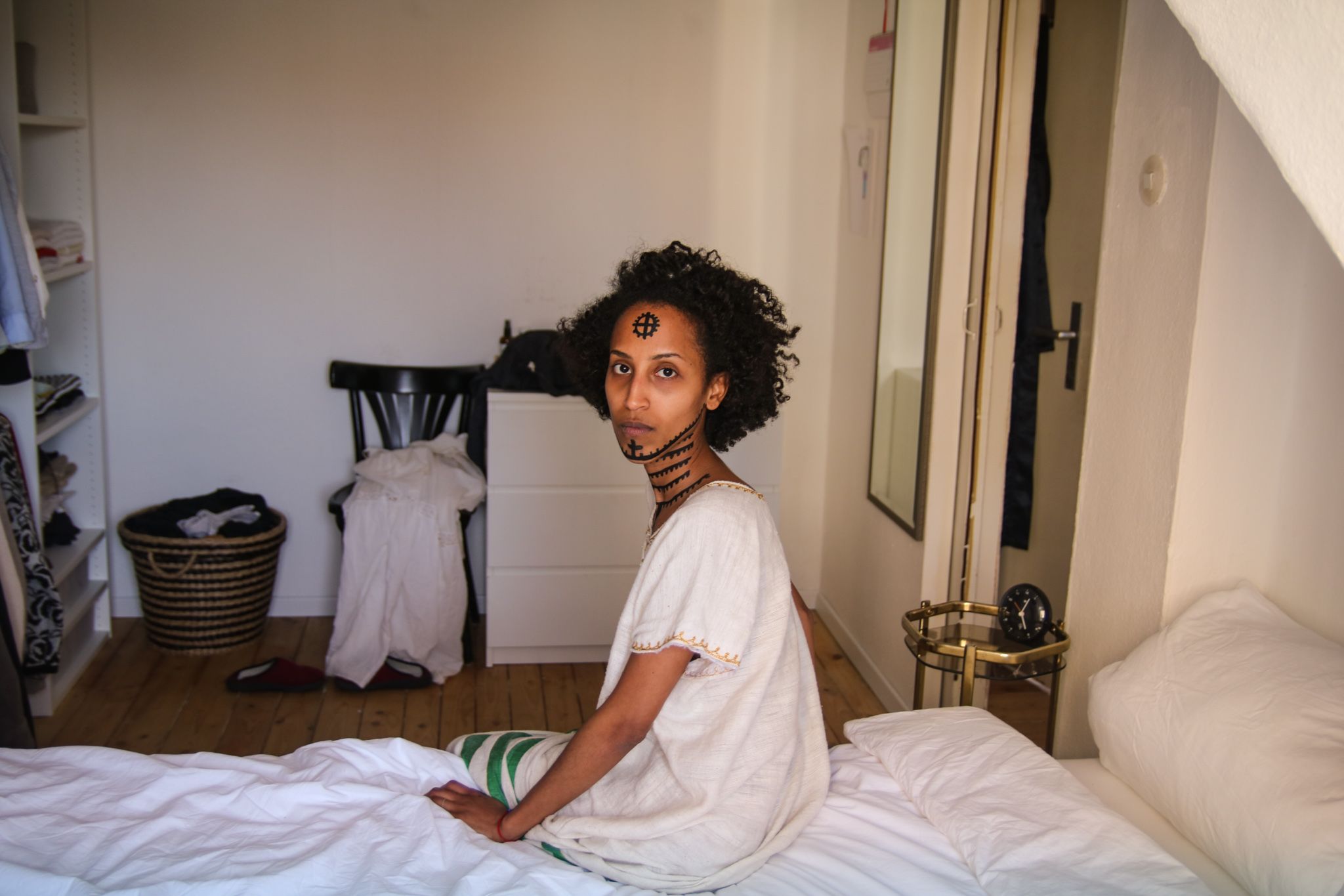 Eine Frau sitzt auf der Kante eines weiß bezogenen Bettes. Ihr Oberkörper und Kopf sind seitlich nach links hinter sich gedreht. Sie blickt nach oben, direkt in die Kamera. Ihr Hals, Kinn und ihre Stirn sind mit äthiopischen Tätowierungen bemalt.