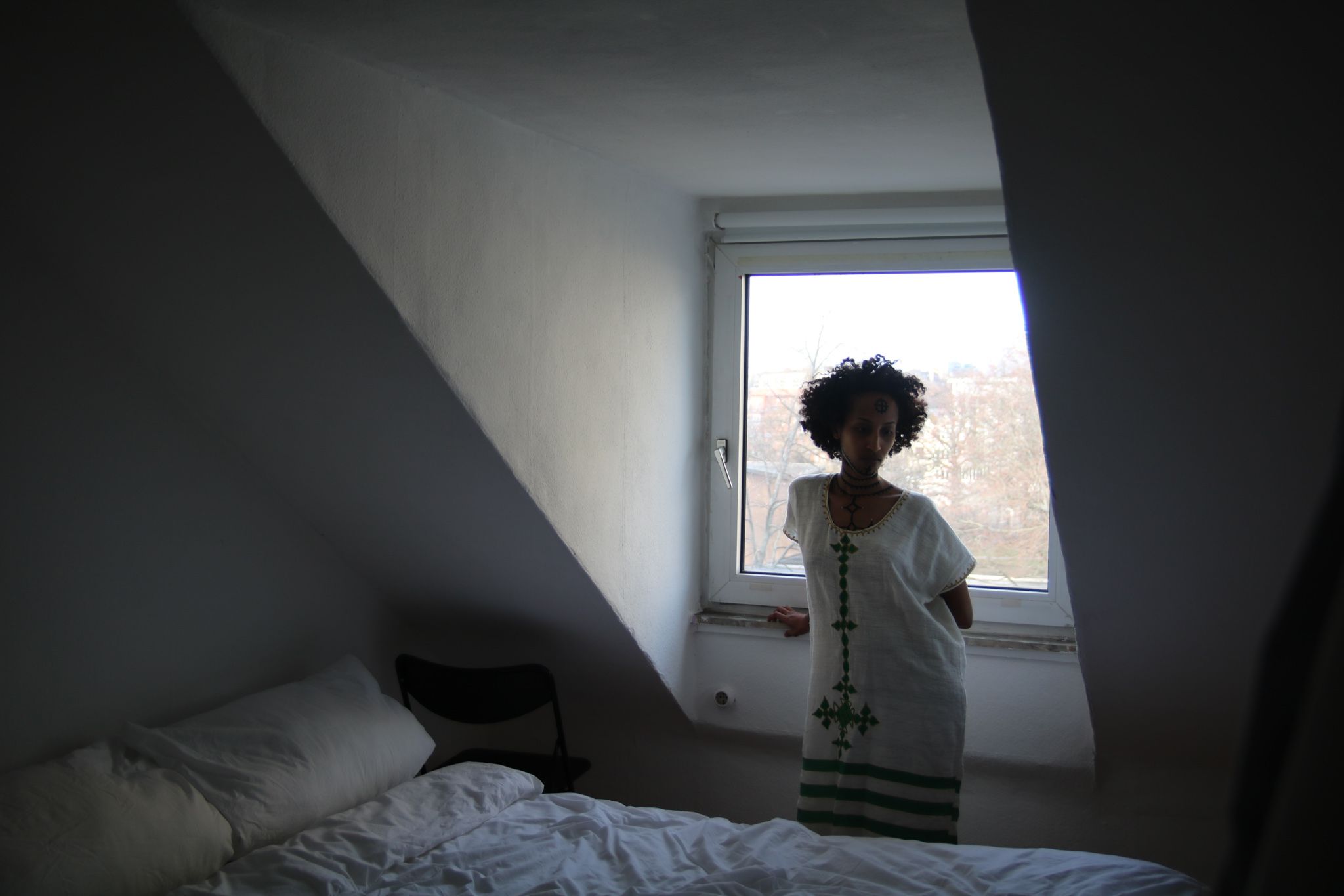 Eine Frau steht im Hintergrund des Bildes vor einem Fenster mit Dachschrägen. Ihr Körper ist dem Raum zugewandt. Vor ihr steht ein Bett. Sie trägt ein weites, weißes Kleid mit grünen Ornamenten. Oberkörper, Hals und Gesicht sind mit Tätowierungen bemalt