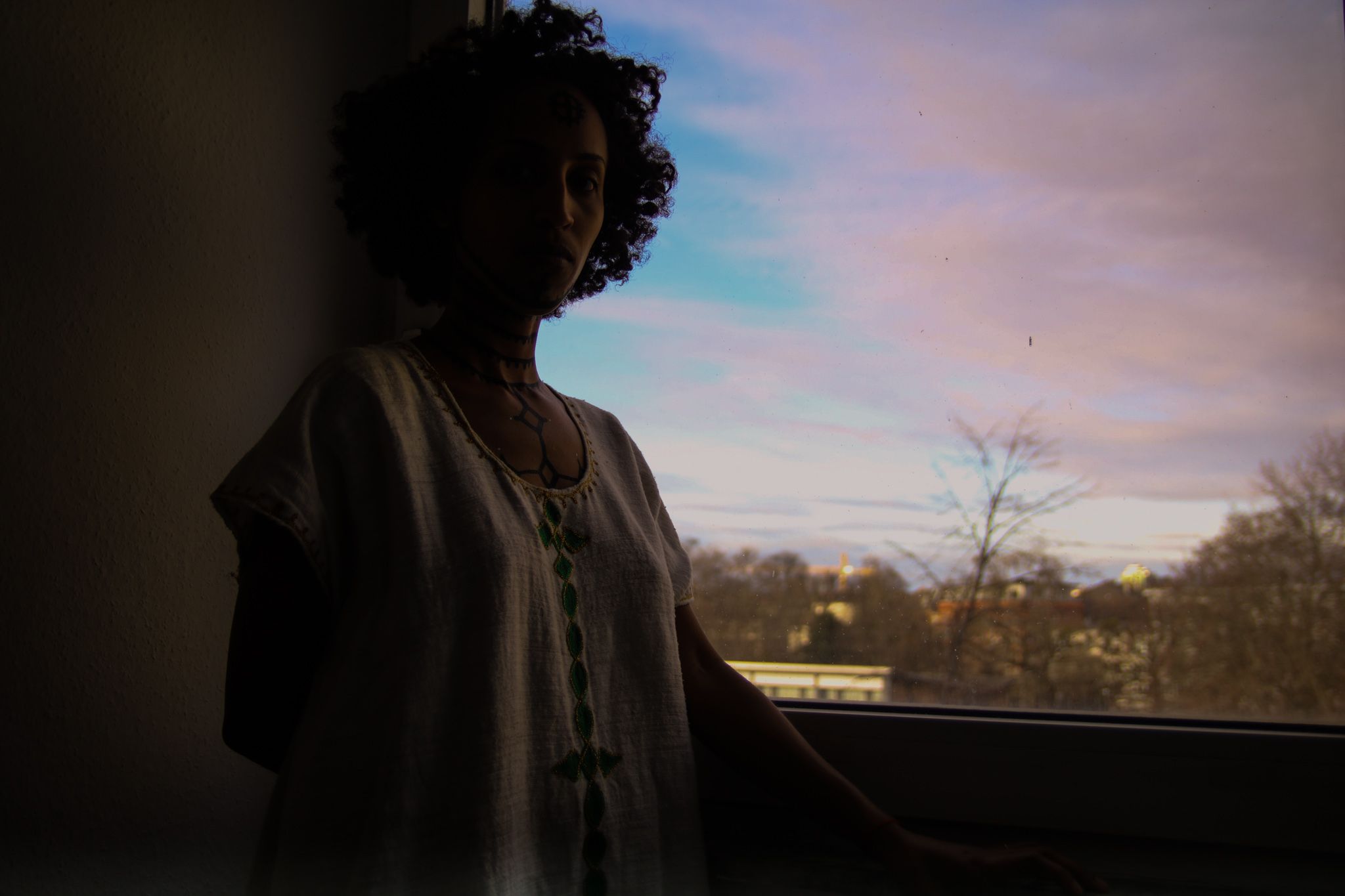 Eine Frau steht am Fenster. Der Abendhimmel ist blau-rosa. Frau ist schemenhaft von der Hüfte aufwärts zu sehen. Sie trägt ein Kleid mit horizontal verlaufenden Ornamenten. Ihr Oberkörper und Hals sind mit äthiopischen Tätowierungen bemalt.