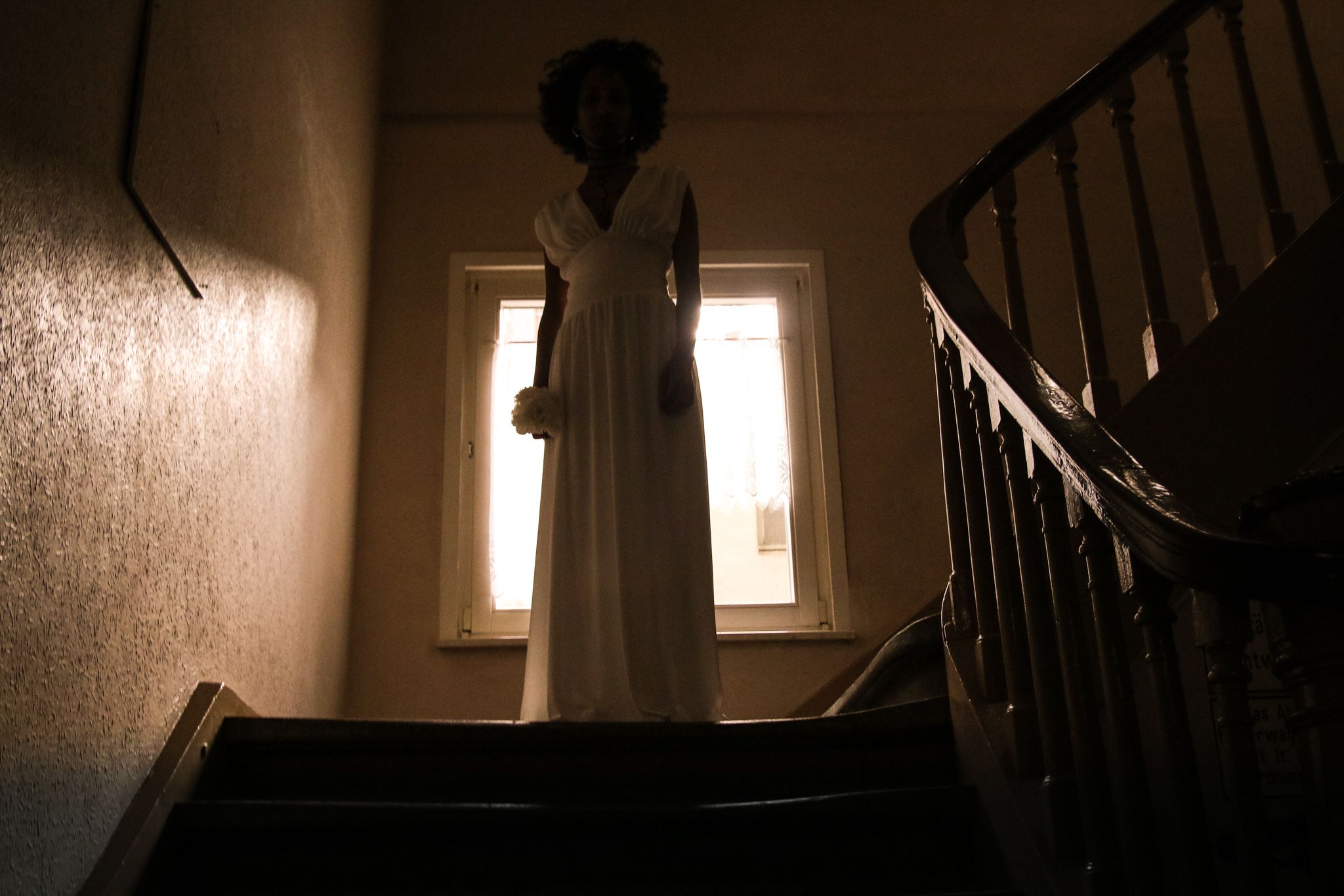 Nach unten gewandt, steht auf dem ersten Treppenabsatz eine weiß gekleidete Frau. Hinter ihr fällt leicht das Licht ins Treppenhaus. In ihrer rechten Hand hält sie eine weiße Blume. Ihr Körper ist der Kamera zugewandt.