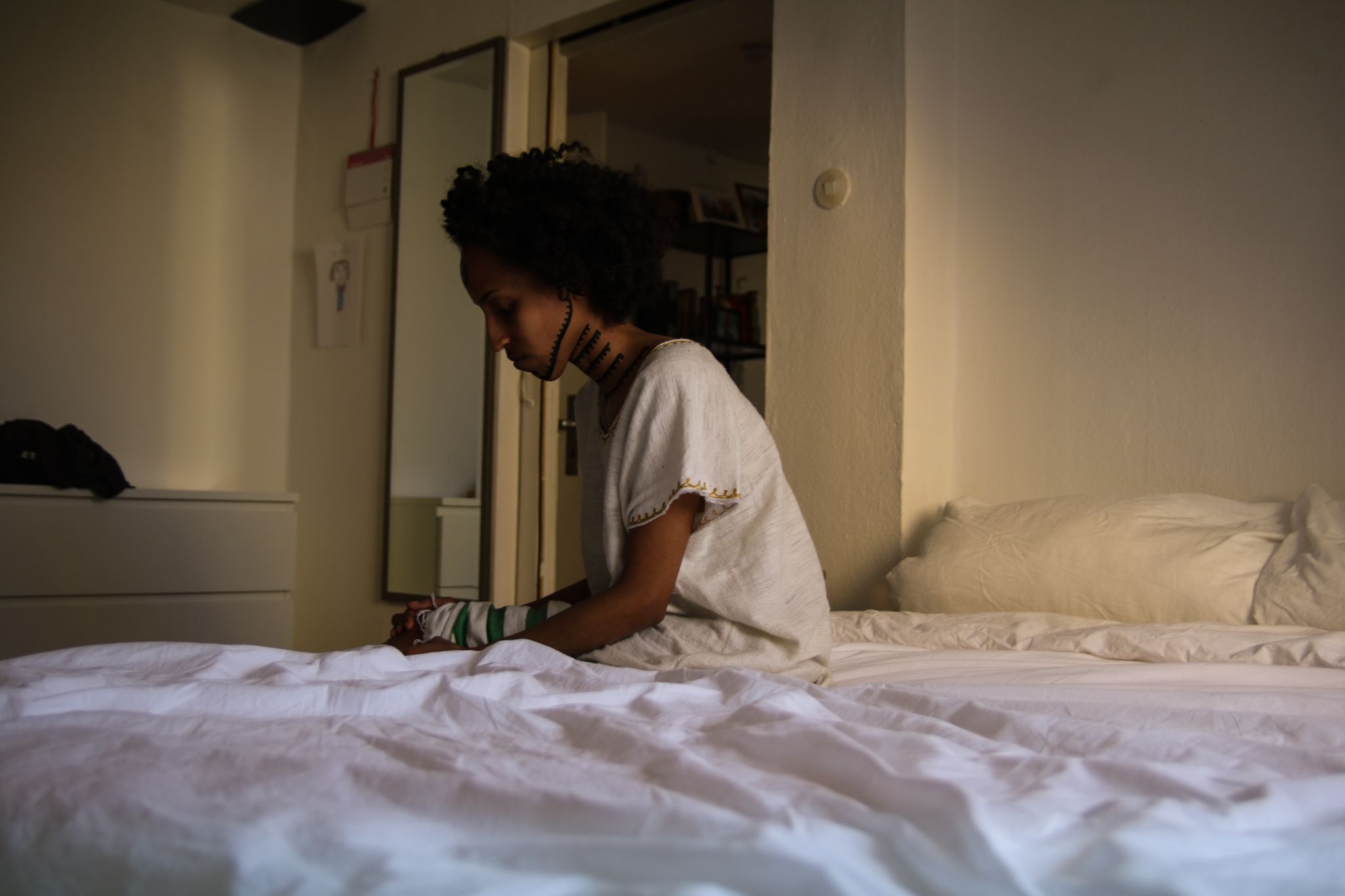 Eine Frau sitzt auf der Kante eines Bettes. Ihr Oberkörper ist von der linken Seite im Profil zu sehen. Sie blickt vor sich nach unten. Ihre Hände umschließen ihre Knie. Am Hals und Kiefer ist sie mit traditionellen äthiopischen Tätowierungen bemalt.