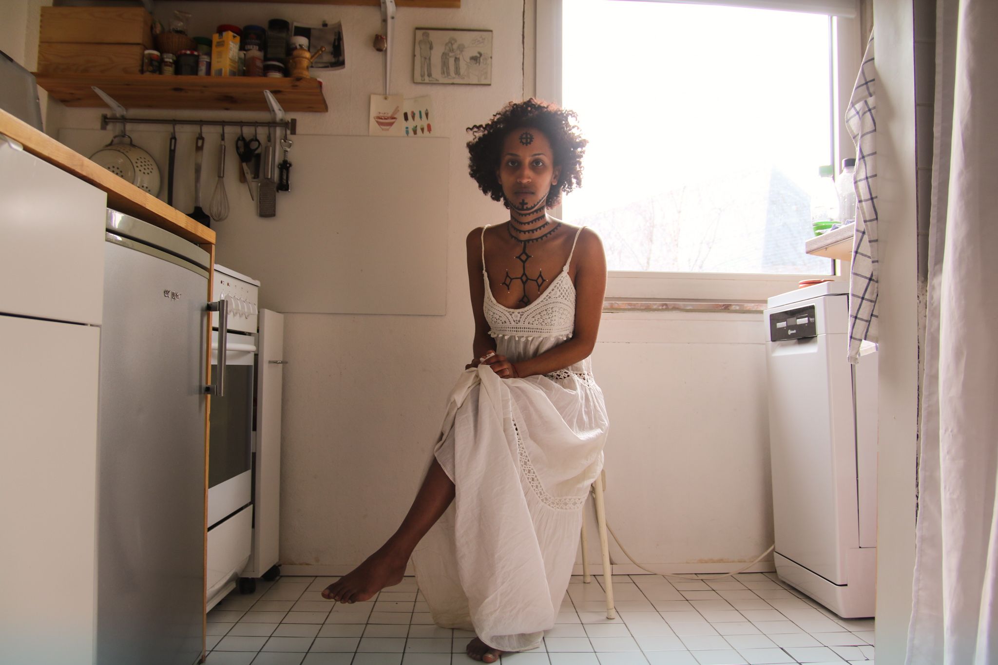 Mittig im Bild in einer hellen Küche sitzt eine Frau mit dem Rücken vor der Küchenwand. Rechts hinter ihr ist ein Fenster. Die Frau trägt ein weißes Kleid. Ihr Brustkorb, Hals und Gesicht sind mit traditionellen äthiopischen Tätowierungen bemalt.