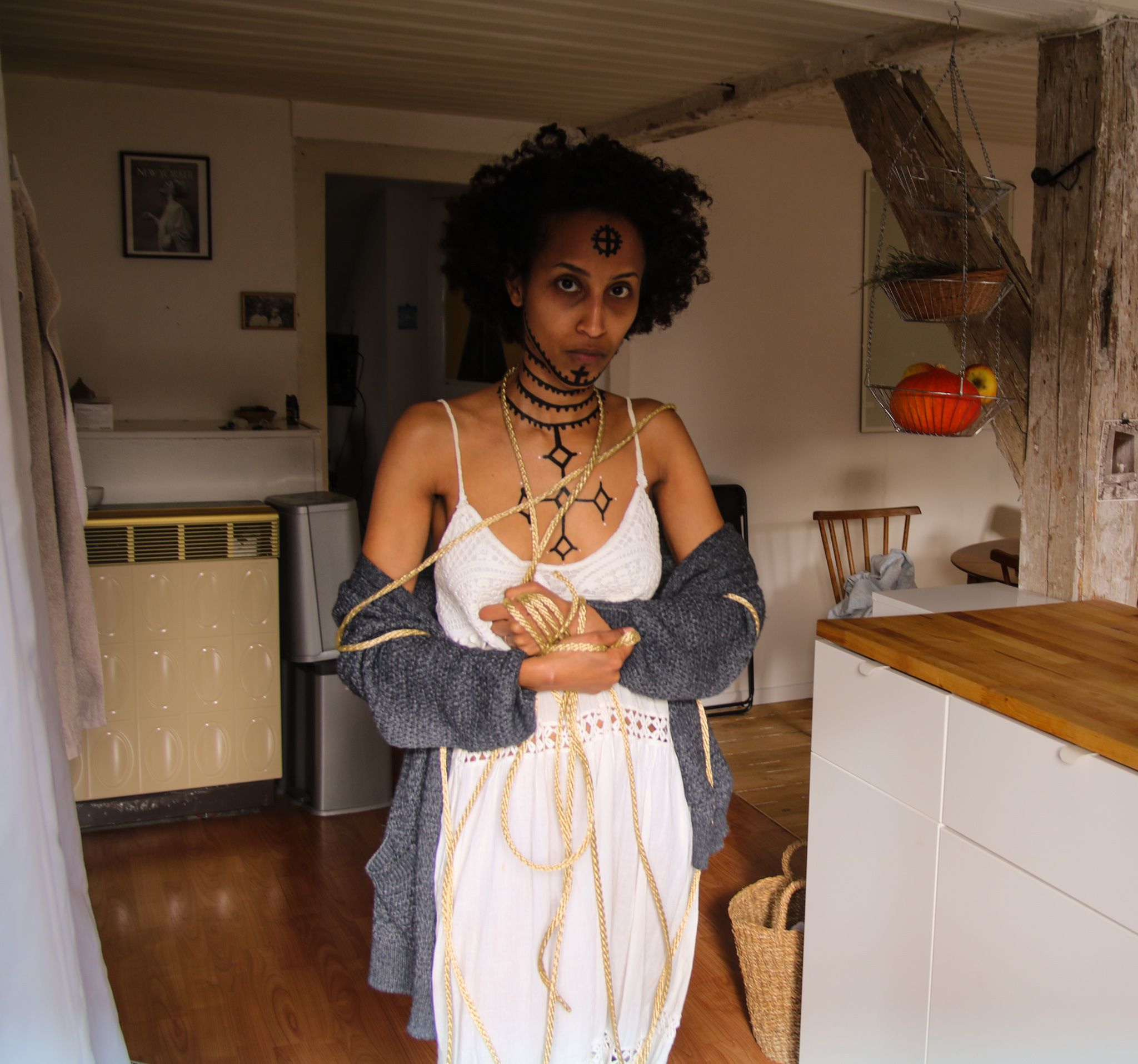Im Bildmittelpunkt steht eine Frau mit Blick in die Kamera. Ihr Brustkorb, Hals und Gesicht sind mit äthiopischen Tätowierungen bemalt. Sie hält ihre Arme leicht vor ihrem Bauch verschränkt. Um ihren Körper und Hände ist ein Seil geschlungen.