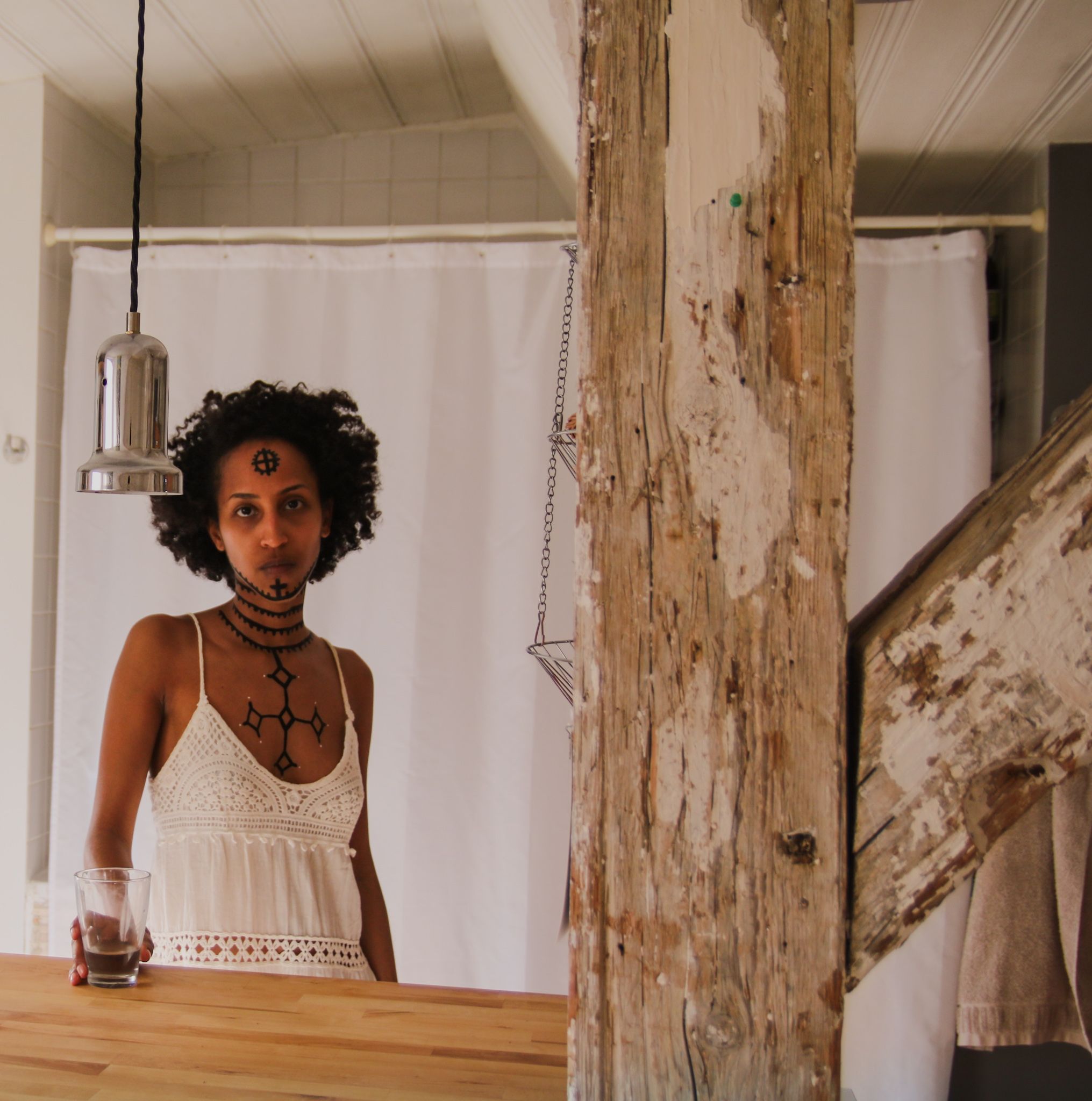 Hinter einem Balken steht eine Frau. Ihr Brustkorb, Hals und Gesicht ist mit äthiopischen Tätowierungen bemalt. Sie hält ein leeres Glas in ihrer rechten Hand, abgestützt auf einer Holzplatte. Sie trägt ein weißes Kleid und blickt direkt in die Kamera.